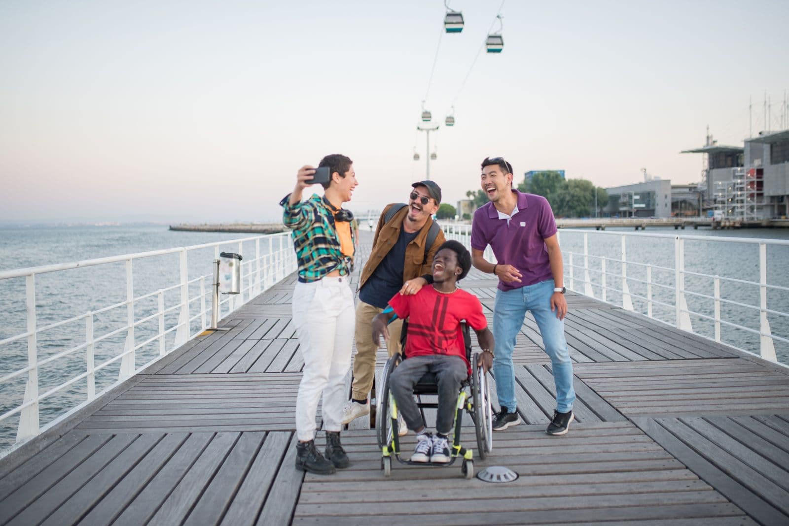 Four joyful adult friends taking a selfie on a waterfront pier, one of them using a wheelchair