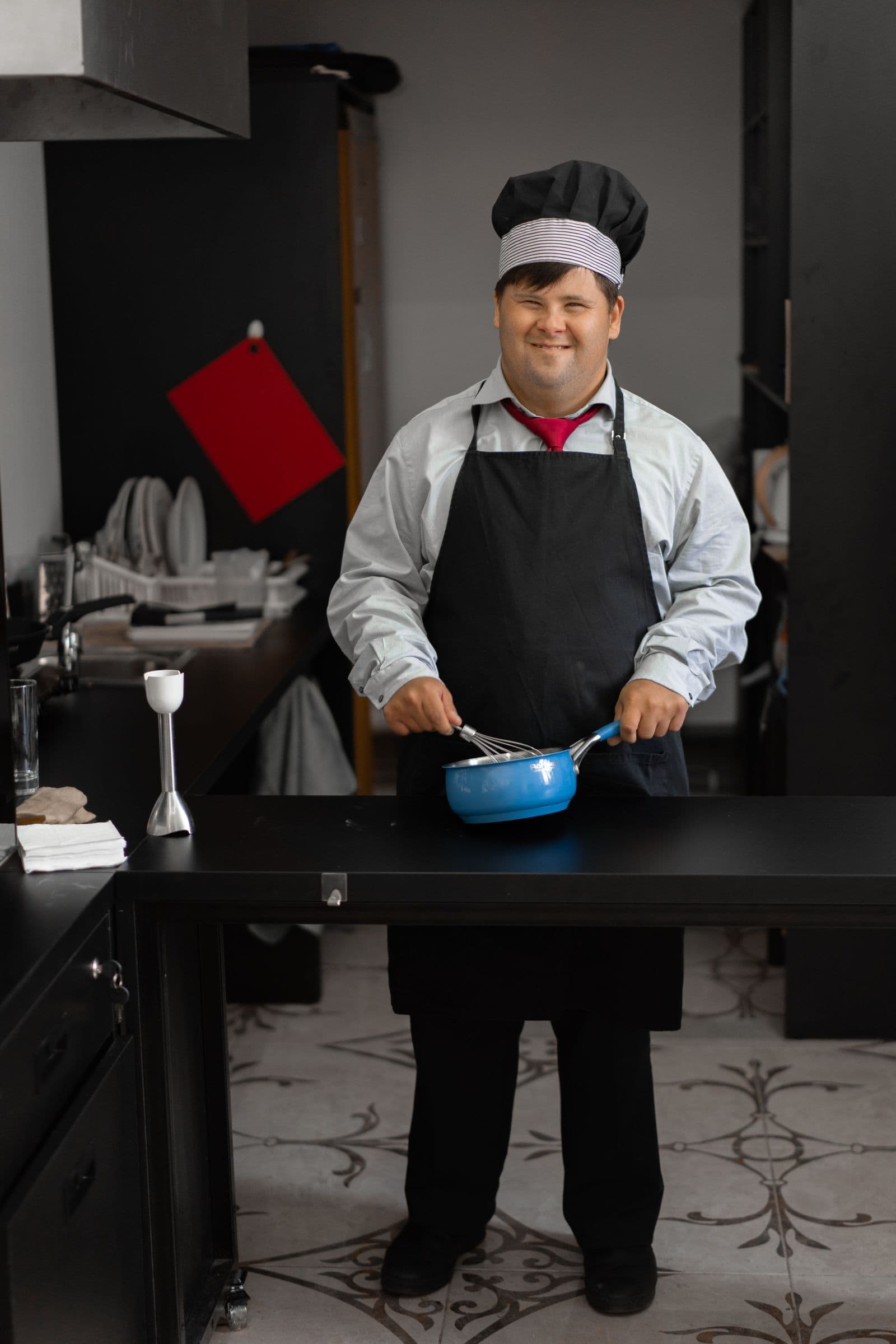 An adult man with Down syndrome, smiling in a chef hat and apron while cooking