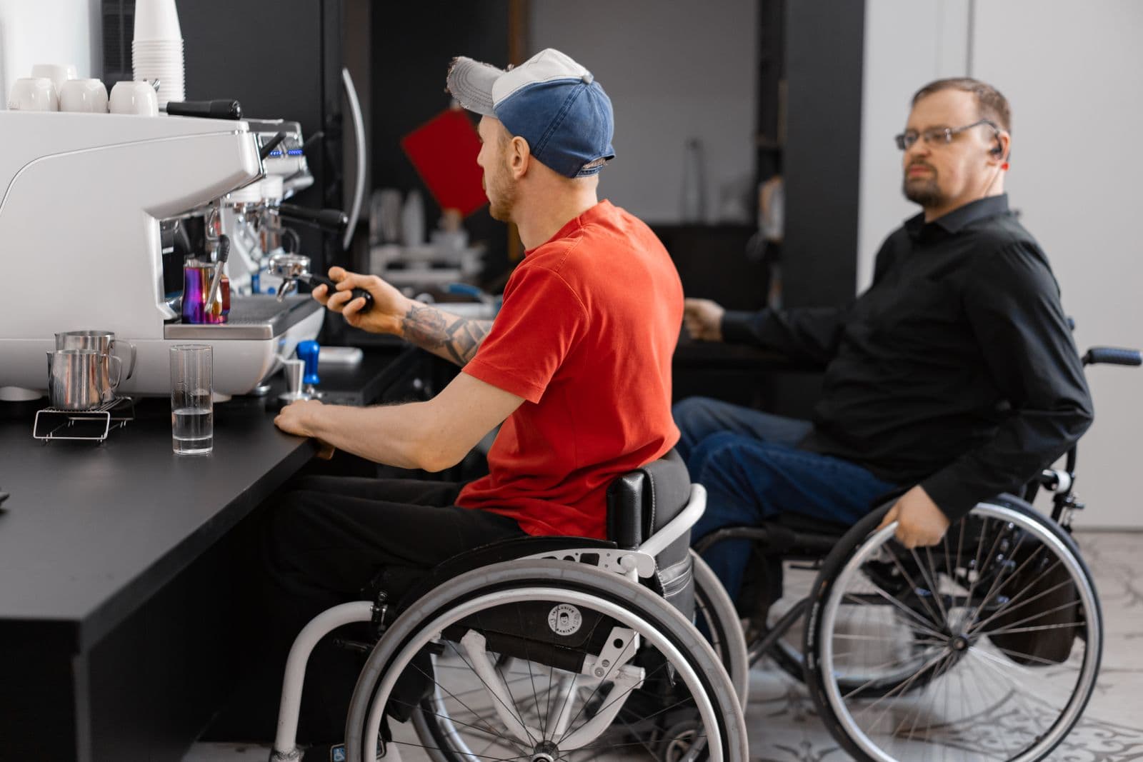 Two adult wheelchair users working together behind an espresso machine in a cafe